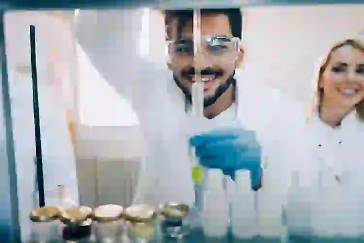 Male lab technician smiling with female lab technician in the background, working with a pipette